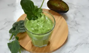 Zielony, kremowy smoothie ze szpinakiem, udekorowane listkiem szpinaku. Obok połówka awokado.