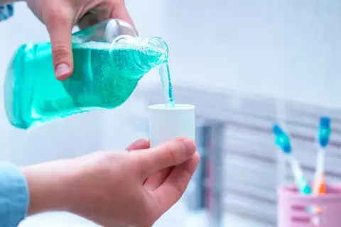 Niebieski płyn do płukania ust nalewany z butelki do małego kubeczka. W tle stoją szczoteczki do zębów.