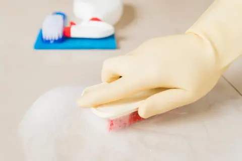 Ręka w żółtej rękawiczce szoruje podłogę szczotką z pianą. Obok leżą akcesoria do sprzątania, w tym butelka z domowym płynem do sprzątania.