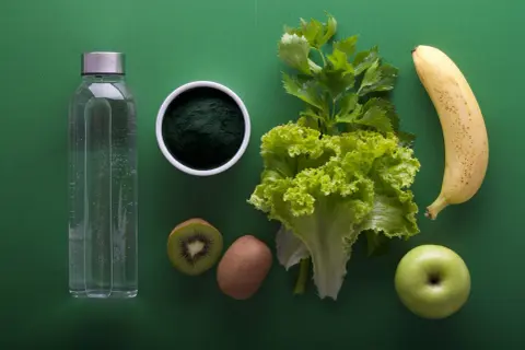 Japońska dieta wodna: butelka wody, spirulina, kiwi, sałata, seler, jabłko i banan na zielonym tle.