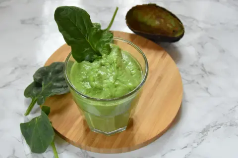 Zielony, kremowy smoothie ze szpinakiem, udekorowane listkiem szpinaku. Obok połówka awokado.