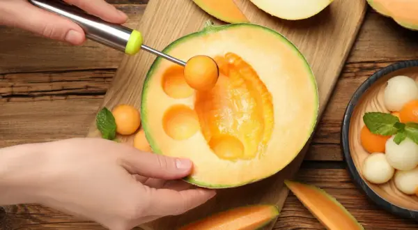 Ręka wydrąża kulki z melona, pokazując, czy melon jest zdrowy. Wokół świeże owoce i listki mięty.