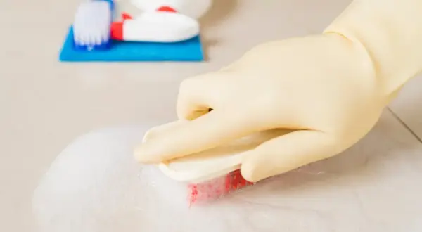 Ręka w żółtej rękawiczce szoruje podłogę szczotką z pianą. Obok leżą akcesoria do sprzątania, w tym butelka z domowym płynem do sprzątania.
