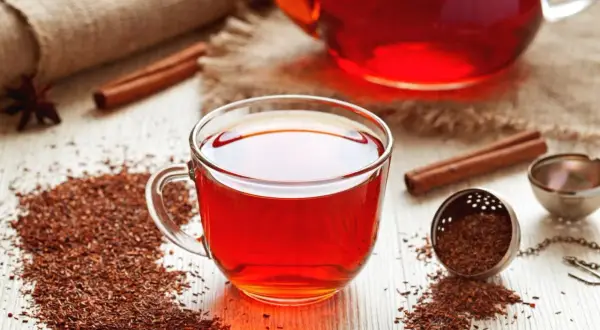 Aromatyczna herbata rooibos, znana ze swoich licznych właściwości, w szklanej filiżance, obok zaparzacza i laski cynamonu.