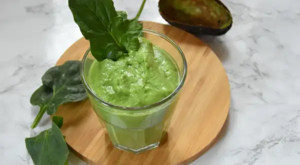 Zielony, kremowy smoothie ze szpinakiem, udekorowane listkiem szpinaku. Obok połówka awokado.
