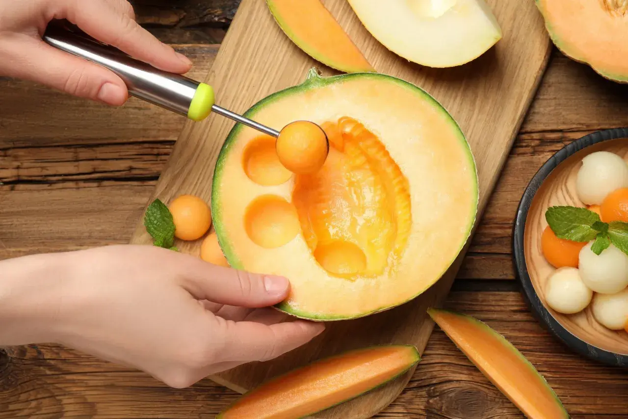 Ręka wydrąża kulki z melona, pokazując, czy melon jest zdrowy. Wokół świeże owoce i listki mięty.