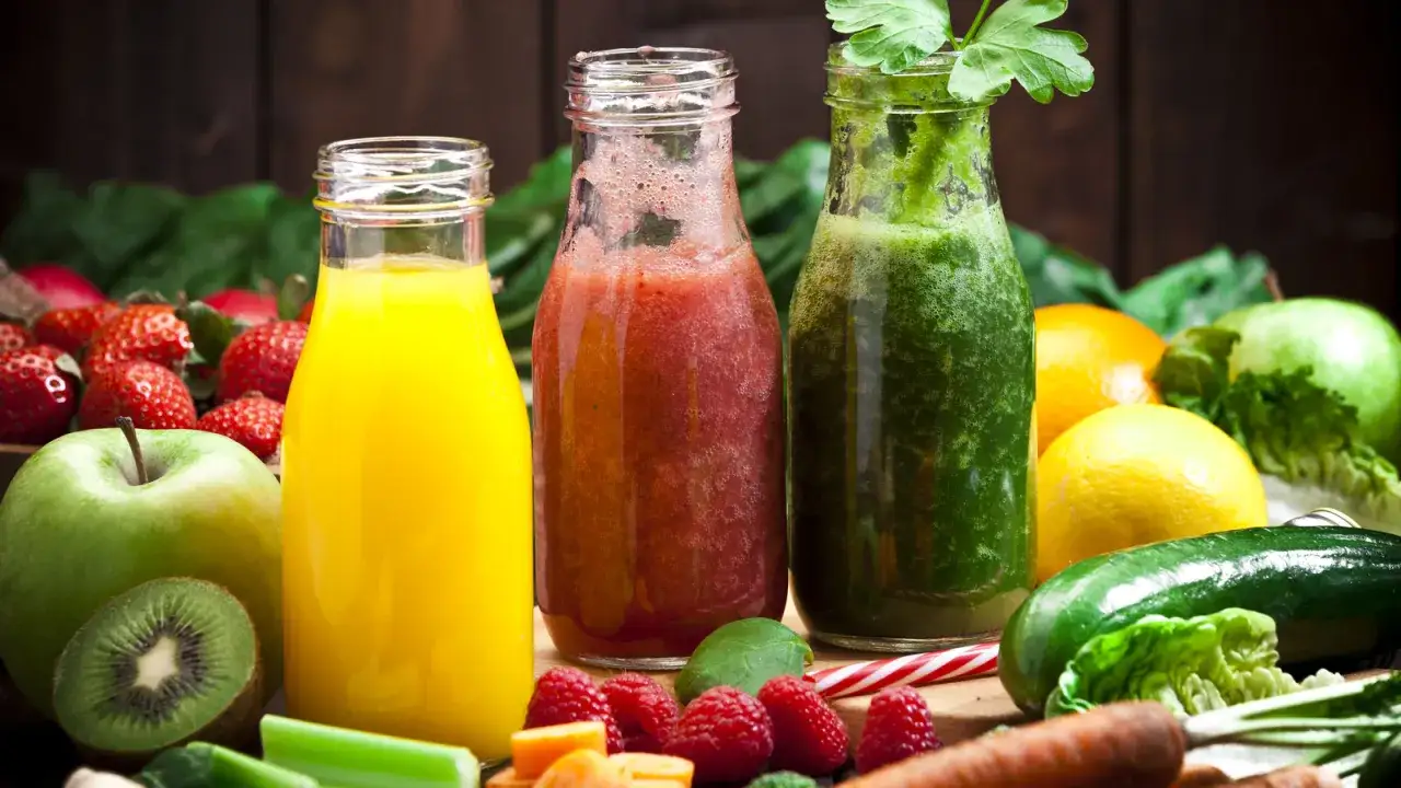 Trzy butelki z kolorowymi sokami: ż&oacute;łtym, czerwonym i zielonym. Obok świeże owoce i warzywa, idealne na detox sokowy.
