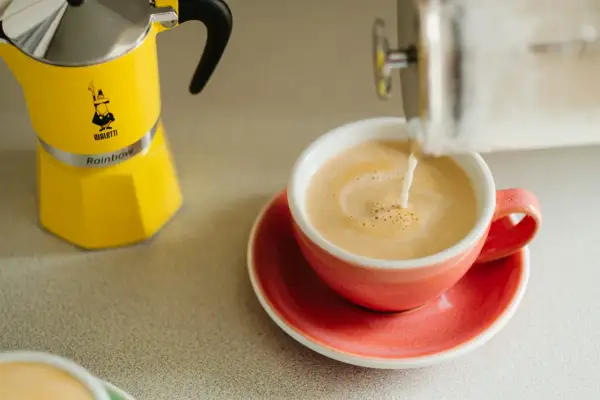 Żółta kawiarka Bialetti obok różowej filiżanki, do której nalewane jest mleko. Idealna kawa z miodem na dobry początek dnia.