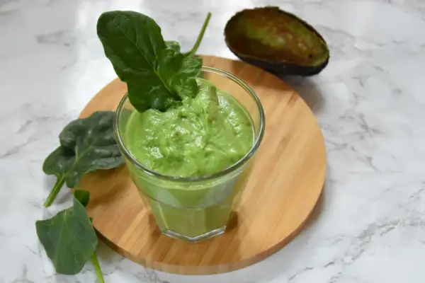 Zielony, kremowy smoothie ze szpinakiem, udekorowane listkiem szpinaku. Obok połówka awokado.